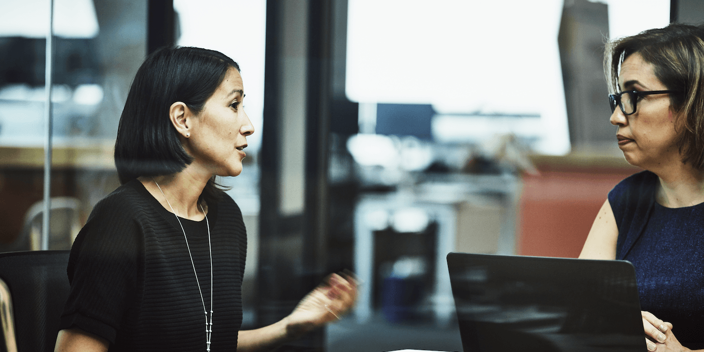 two people sitting in a conference room in an office, one speaking as the other listens