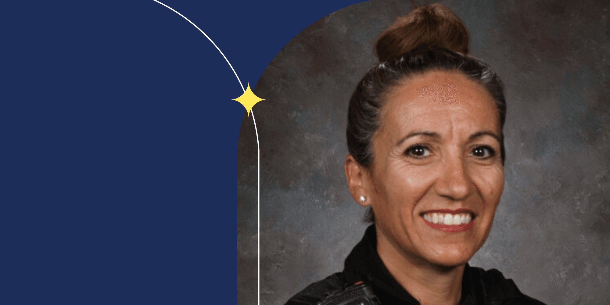 A smiling person facing the camera. They wear their hair in a bun on the top of their head. They also wear earrings, red lipstick, and a black shirt.