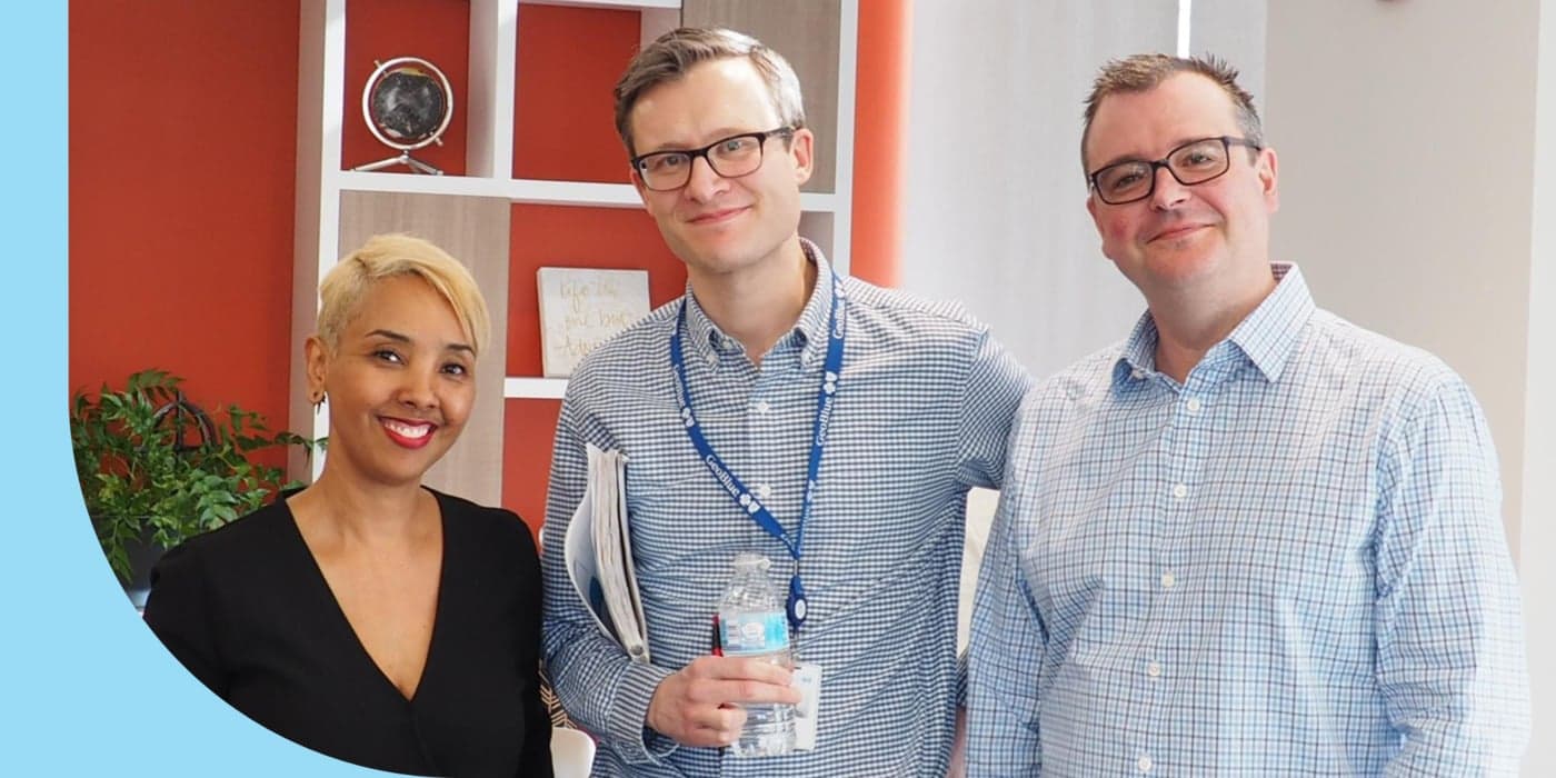 Three people facing the camera and standing in an office space. The person on the far left wears a black blouse and has short blonde hair. The person in the middle wears glasses, a blue button-down shirt, and a lanyard. The person on the far right wears glasses and a button-down shirt. An orange wall can be seen behind them.